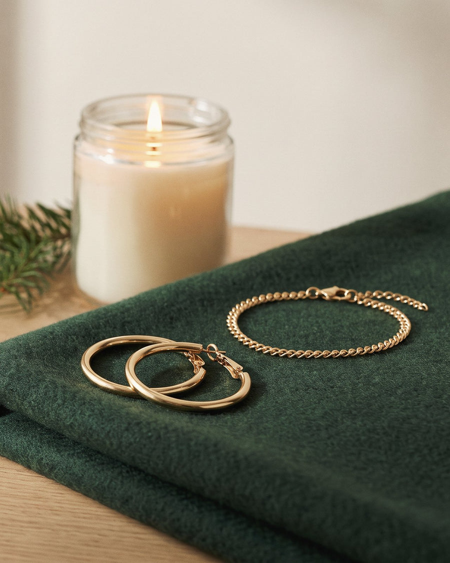 Gold tone hoop earrings and a chain bracelet on a green wool scarf, a candle behind