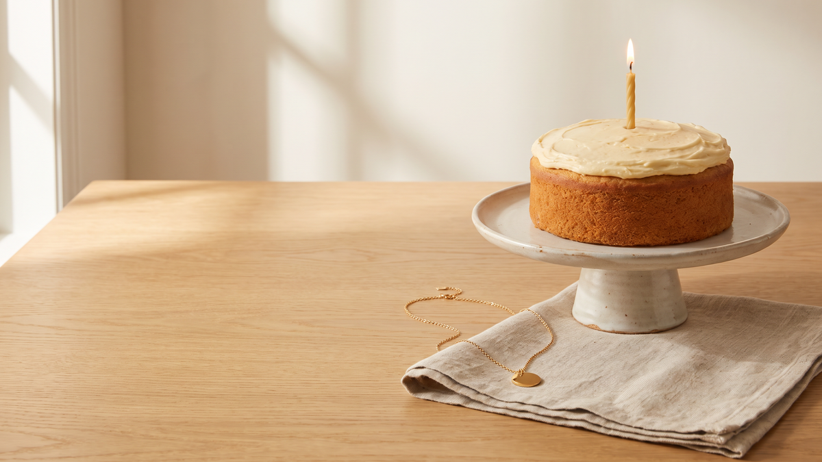 Gold tone pendant necklace on a linen napkin in front of a small cake with one candle