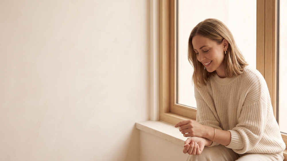 Woman in a cream jumper looking at a slim gold tone chain bracelet on her wrist