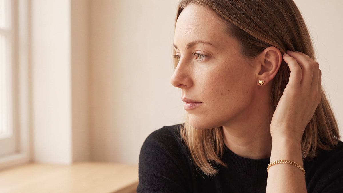 Model in a black jumper tucking her hair behind her ear, wearing gold tone heart stud earrings and a chain bracelet